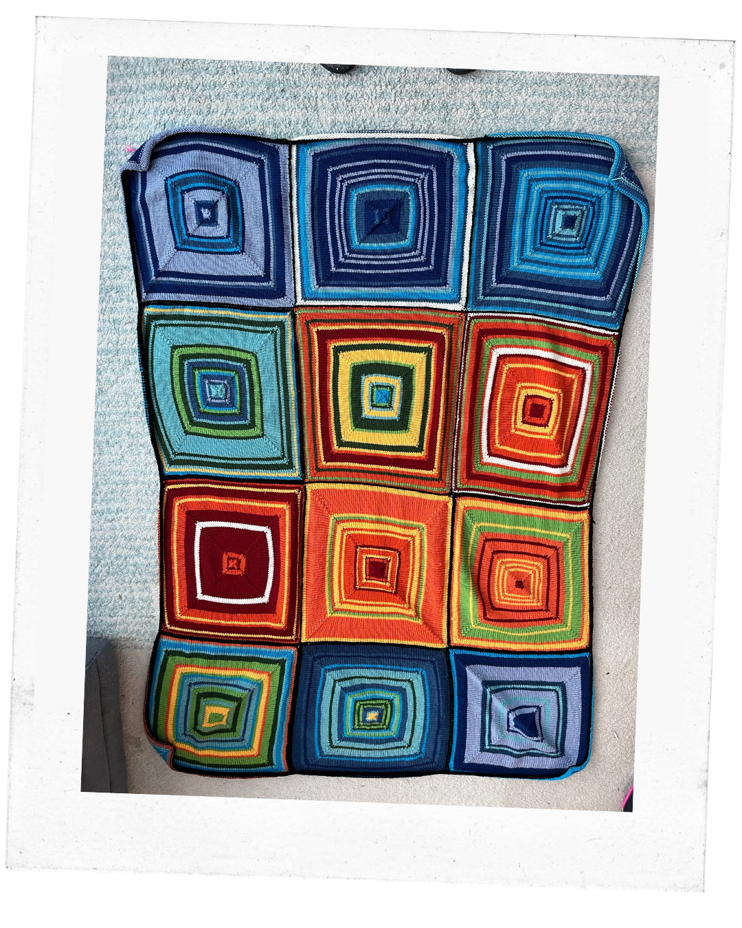 Polaroid of a blanket made up of 12 squares consisting of smaller squares of shades of blues, reds, yellows, green.