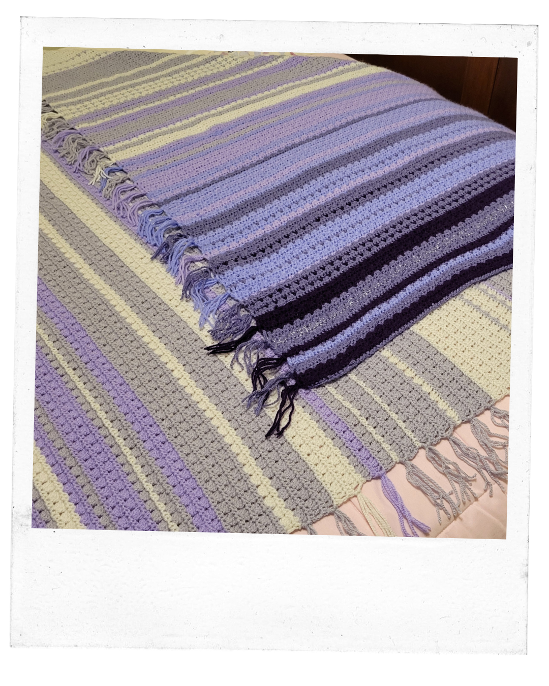 Blanket with lines of light purples, lilac, white and silver, lays folded on a bed. 