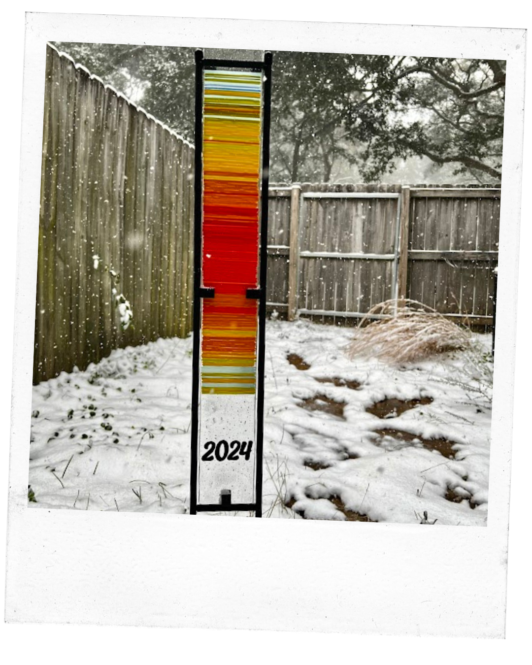 Polaroid of a long piece of glass resembling a scarf, consisting of many different color lines representing temperatures over time, placed in a frame sitting outside surrounded by snow and winter weather.