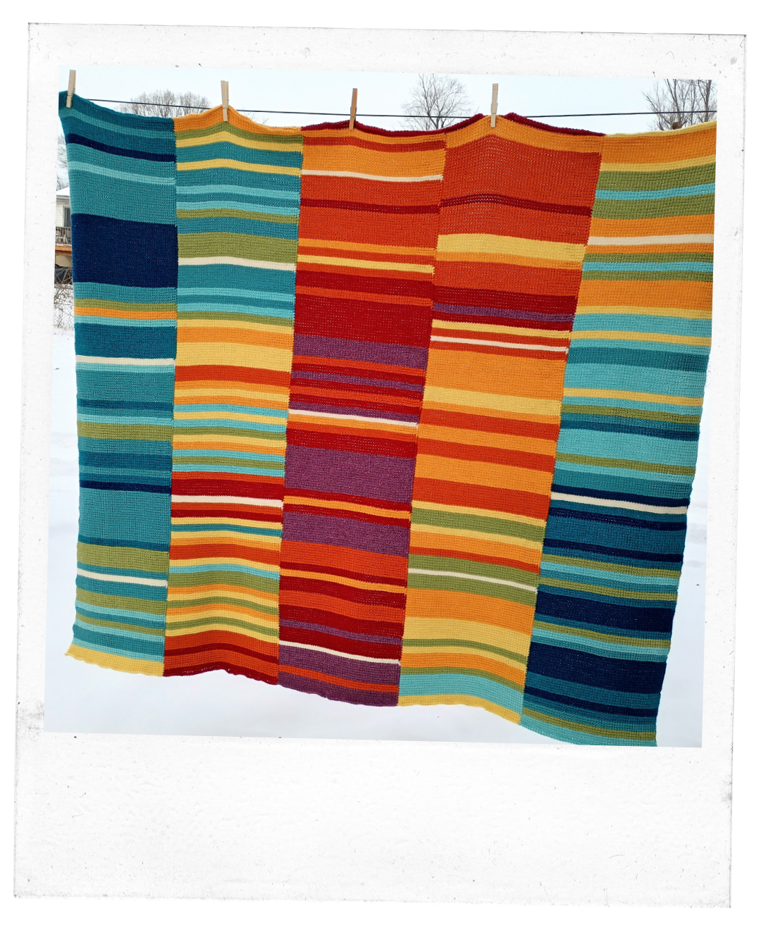 a large crocheted blanket hands from a line with clothes pins, made up of long rows with smaller rows of colors in varying shades of reds, green, blues, yellow and orange. 