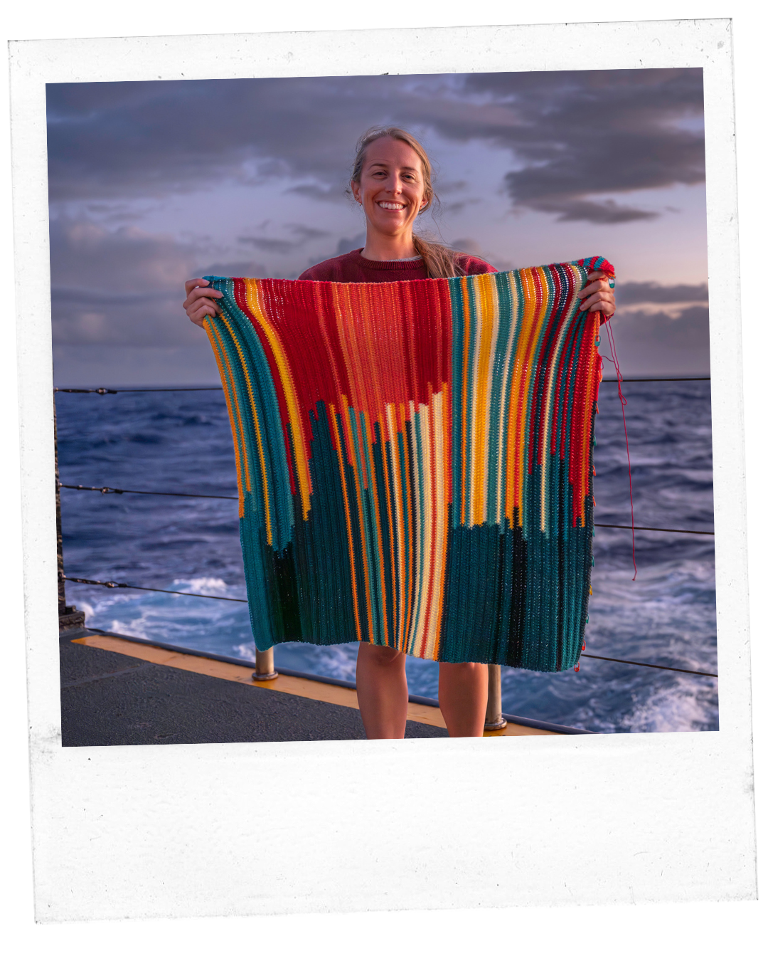 Jessica Gwinn standing in front of the ocean holding the Bathymetry tapestry of lines of pinks, yellows, blues, crocheted to show each day at sea and the sea depth the temperature is from. 