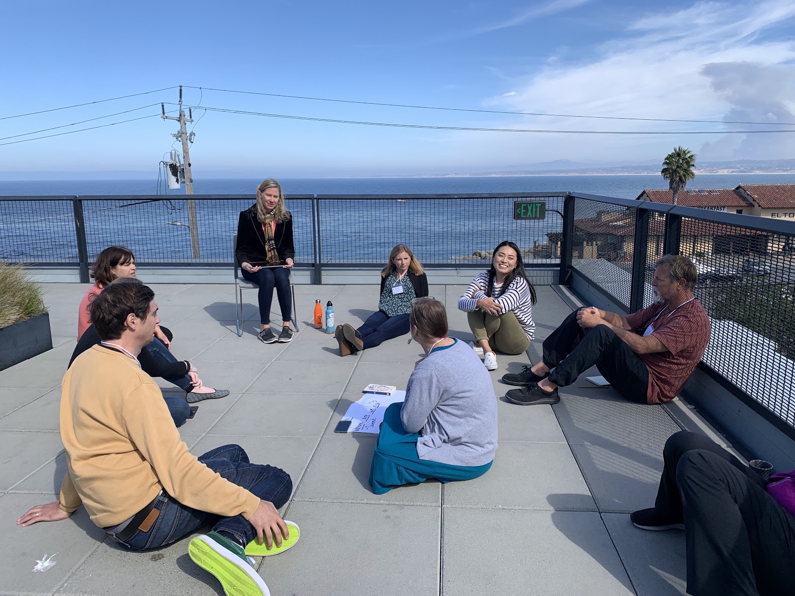 A group of people sit in a circle on a grey patio overlooking a blue bay