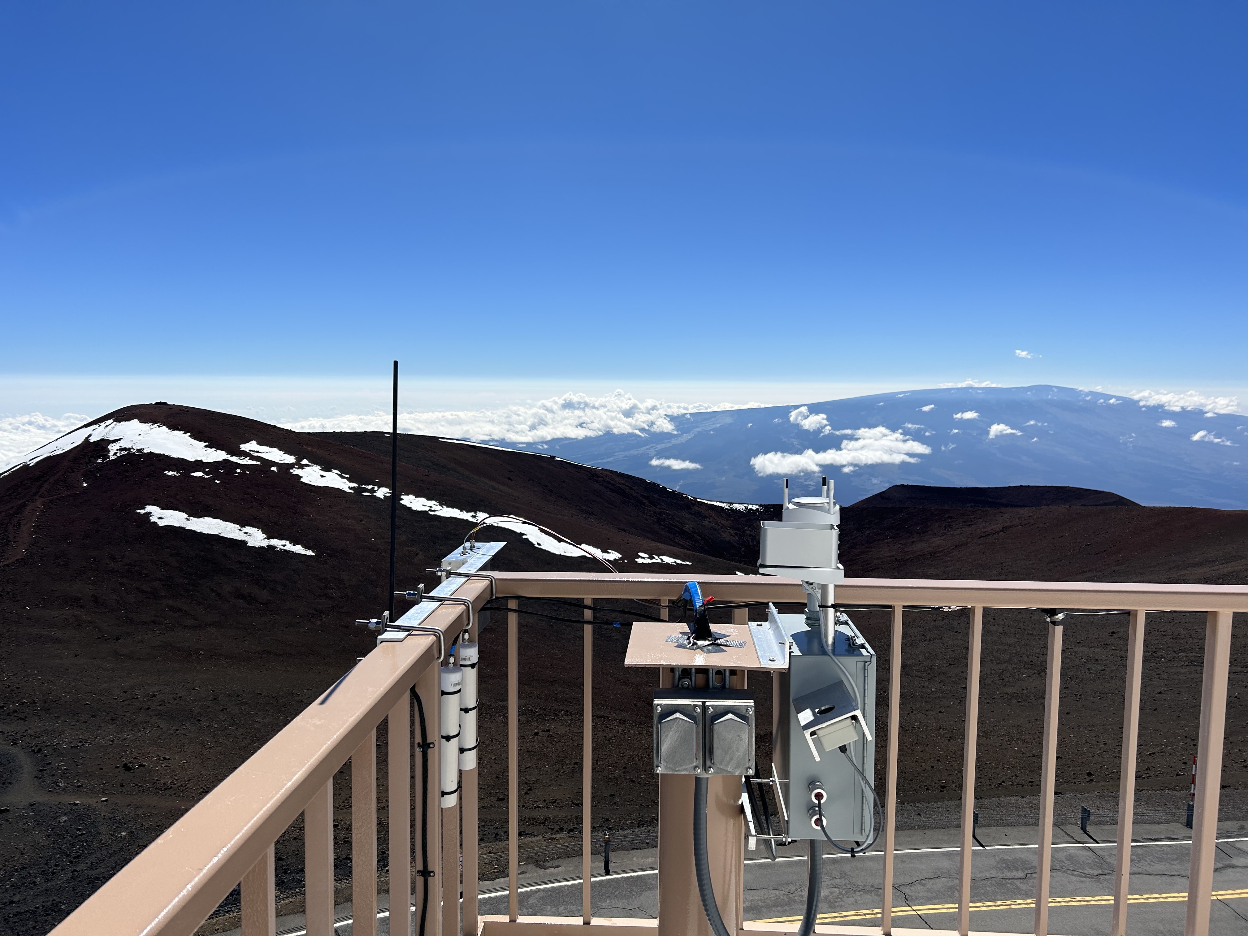 A rectangular tube is attached to the banister of a deck on a building. The deck overlooks mountains with some snow cover and a two lane road. Cloud cover is below the deck height, down in the valleys. 