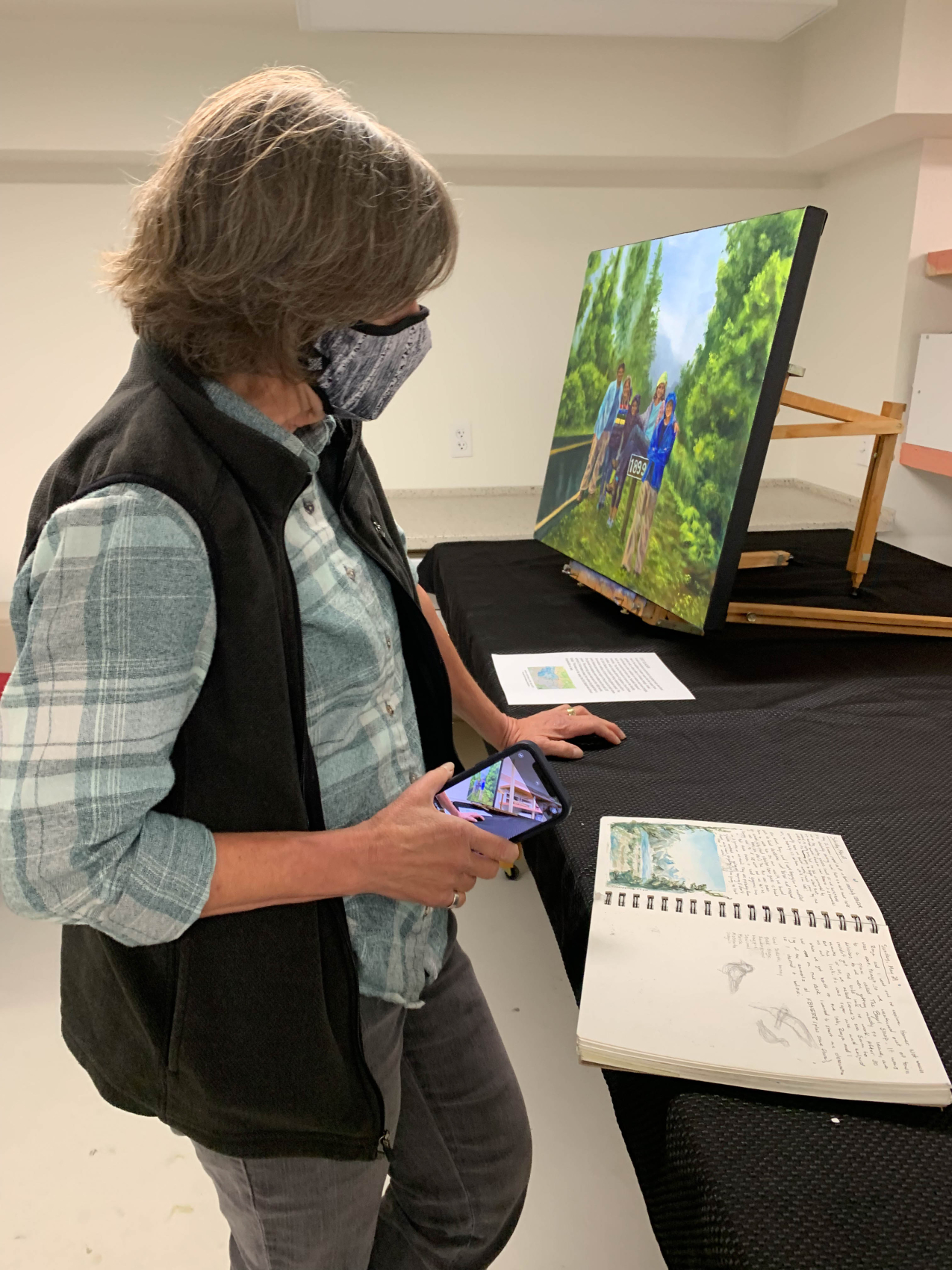 A woman wearing a fabric face mask over her mouth and nose looks at an open sketch book, which contains a watercolor painting and handwritten notes. Behind her, there is a painting of a group of people standing alongside the road next to a sign that reads "1899"