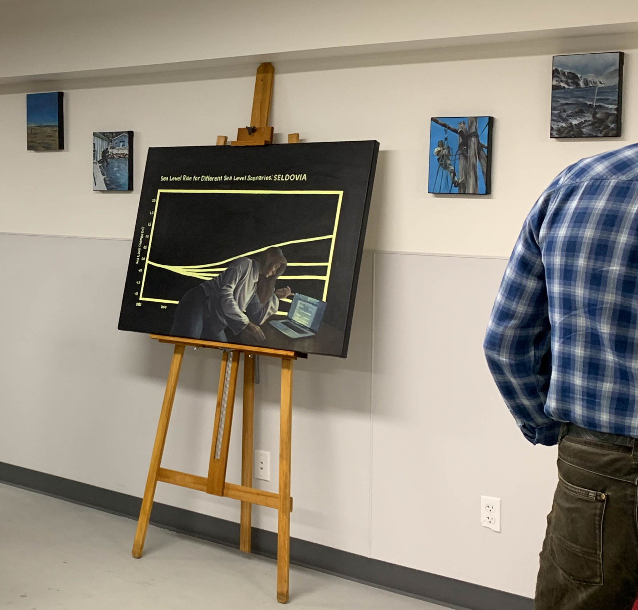 A large painting with two smaller paintings on the wall on either side. The large painting is a data line graph of rising sea level with the title "Sea Level Rise for Different Sea Level Scenarios: Seldovia. A painting of a woman working at a computer is overlaid on the painting of the graph. She appears to be working in coding software on the computer screen.