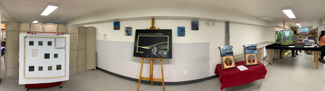 A panorama of Kate's work grouped in modules. Module one: several small paintings are arranged within a sampling quadrat, Module two: a large painting of rising data curve with a woman looking at a computer painting slightly overtop it is surrounded by four smaller paintings. Module three: two paintings atop wooden frames stood up on a table. Module four: another table holds several paintings and a notebook.