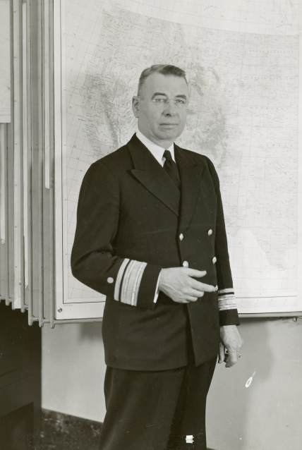 A black and white photo of Rear Admiral Lee Otis Colbert, Director of the U.S. Coast and Geodetic Survey, stands in front of a large map wearing his dress blues.
