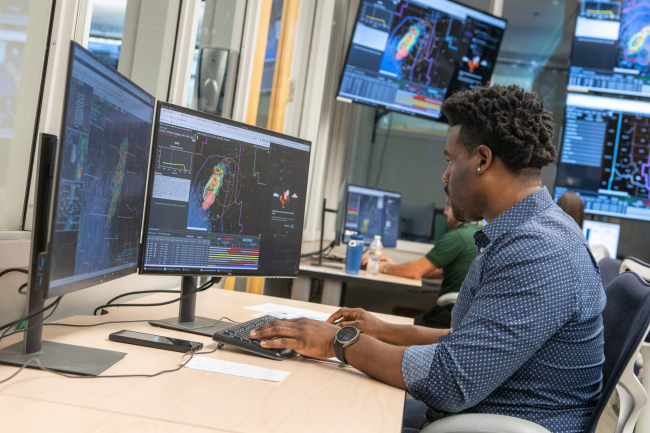A man sits at an office desk and is typing on a keyboard. The two screens in front of him show radar maps and data sets. In the background are more screens on the wall showing additional radar maps. 
