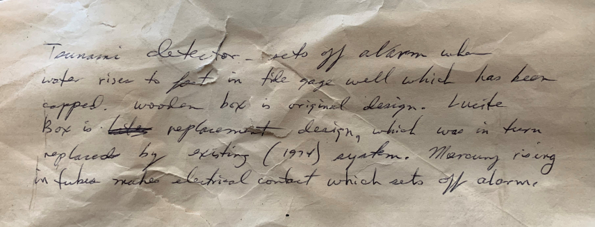 Photo of a handwritten note that was found with the tsunami detector. The note reads, “Tsunami detector sets off alarm when water rises too fast in tide gauge well which has been capped. Wooden box is original design. Lucite box is replacement design, which was in turn replaced by existing (1974) system. Mercury rising in tubes makes electrical contact which sets off alarm.”