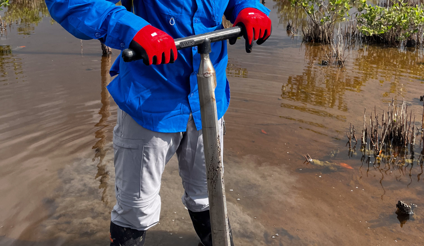Jonah stands in a wetland with muddied water coming midway up his knee-high boots. He pushes down on a meter-long soil corer and looks at the camera.