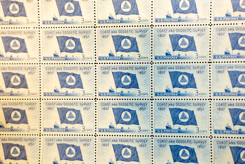 Close up of a sheet of 1957 U.S. Coast and Geodetic Survey 150th anniversary stamps. The stamps feature the USC&GS flag (a red triangle enclosed by a white circle on a blue field) and three ships. They bear the words, "COAST AND GEODETIC SURVEY 1807-1957 U.S. Postage 3 CENTS"