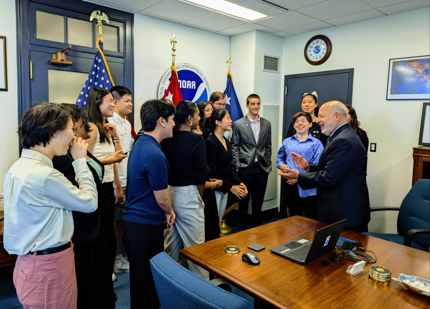 Students smile and laugh while conversing with Dr. Rick Spinrad, NOAA Administrator, in his office. They are standing next to a wooden table and in front of a large NOAA logo and flags.