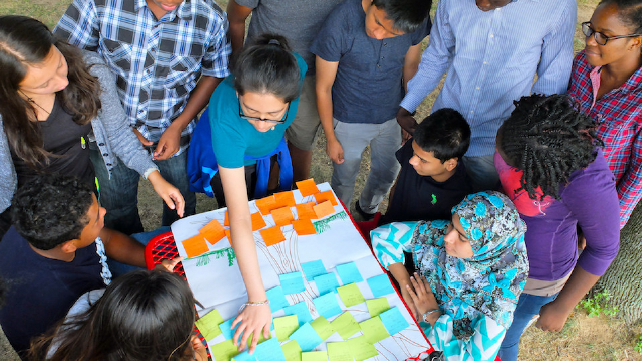 An overhead photo of students gathered around a poster with sticky notes of action items (not readable).