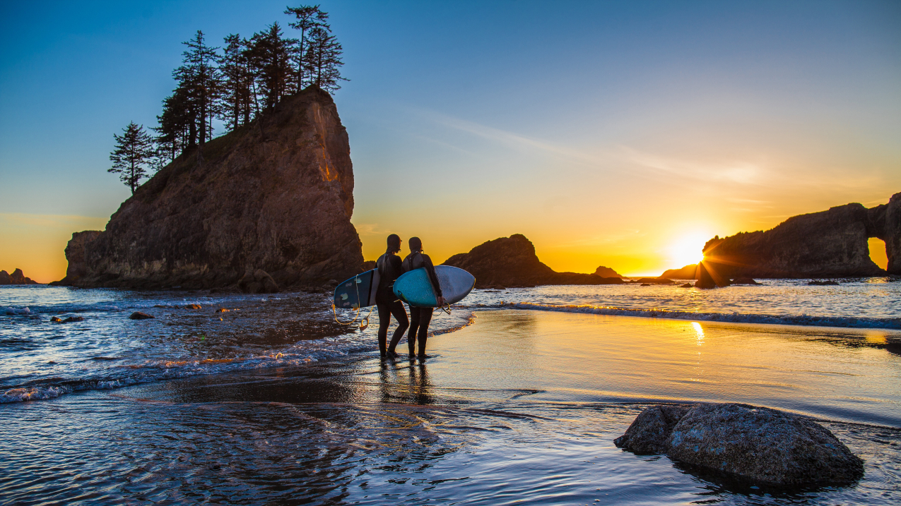 A pair of surfers walk on the shore during sunset in Olympic Coast National Marine Sanctuary