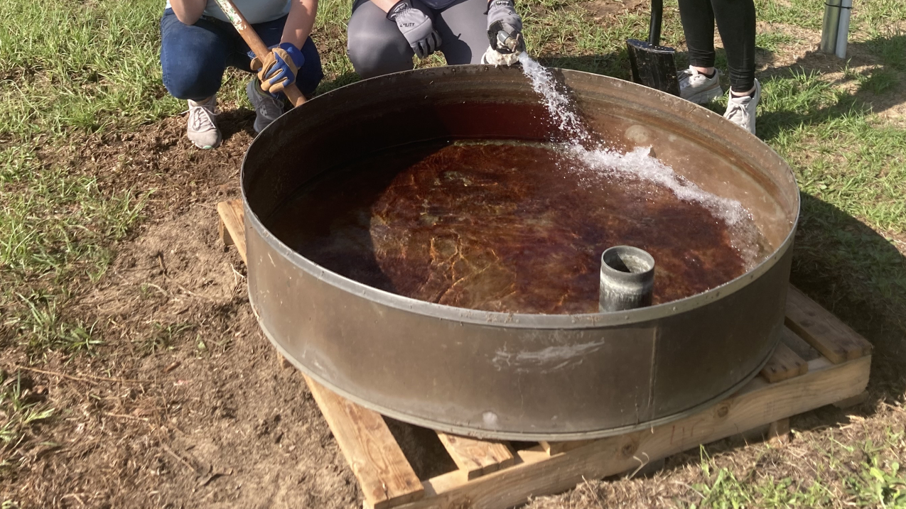 Three women and a large steel pool of water the size of a kiddie pool in front of them surrounded by a flat rural landscape.  Gabby is holding a pickaxe, the woman in the center holds a hose that is filling the pool, and the other holds a shovel. All three smile at the camera.