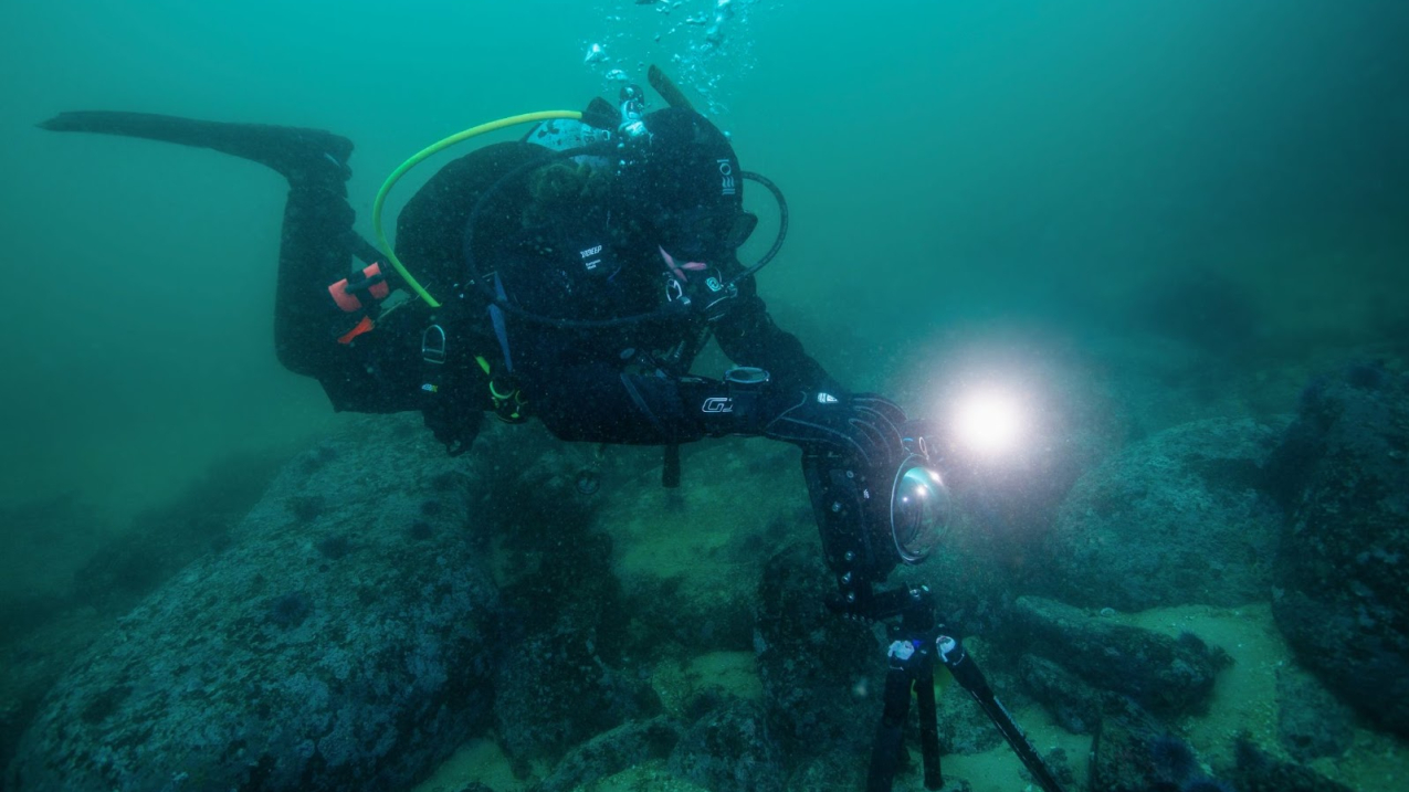 Gracia SCUBA diving. She holds onto a large camera on a tripod that is placed on the ocean floor. The camera's flash is going off.