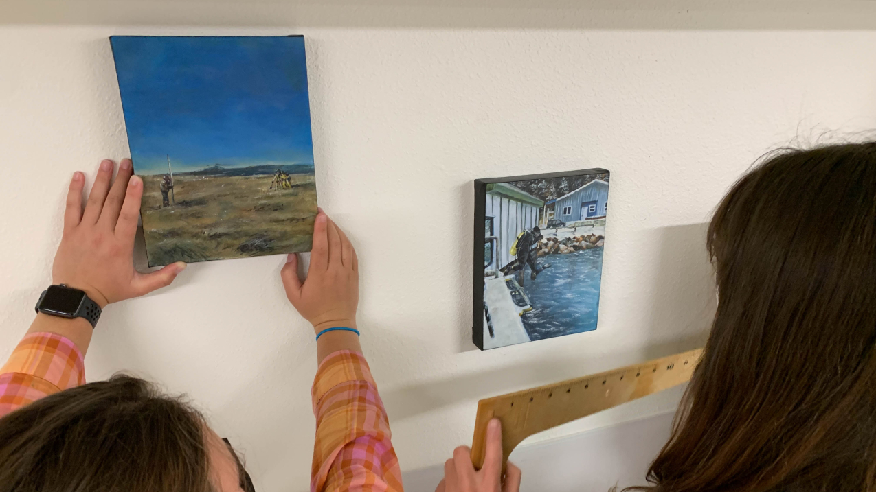 Kate holds a ruler as her sister places a painting. The left painting shows someone holding a pole that extends several feet above them while another person looks through a surveying tripod in a coastal area. The second painting shows a scuba diver mid-jump from a boat docked near the shore.