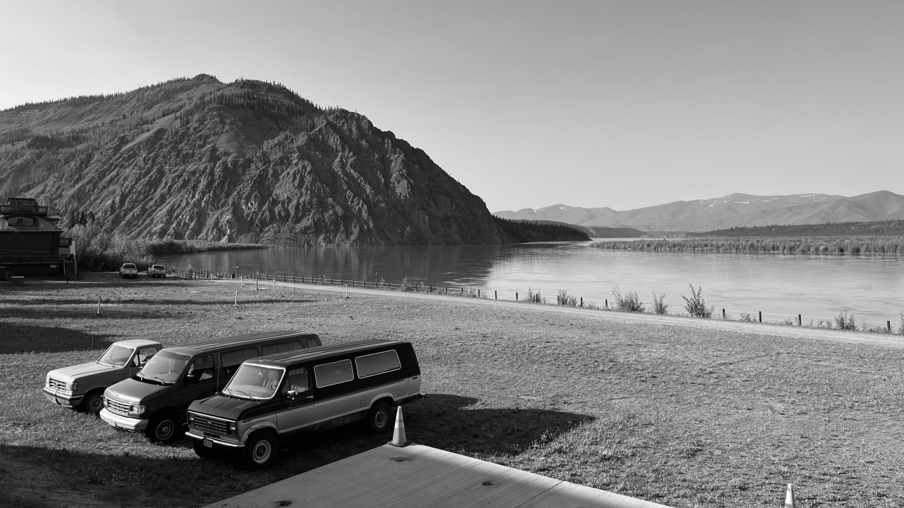 A black and white photo. A nearly empty, grassy parking lot abuts against a wide, calm river. The river bends against a rocky mountain dotted with evergreens and flows into the horizon towards a distant mountain range. A truck and two vans are tucked into the left side of the parking lot, and lend the photo a sense of being taken out of time because they are in good condition, but with a decades old style.