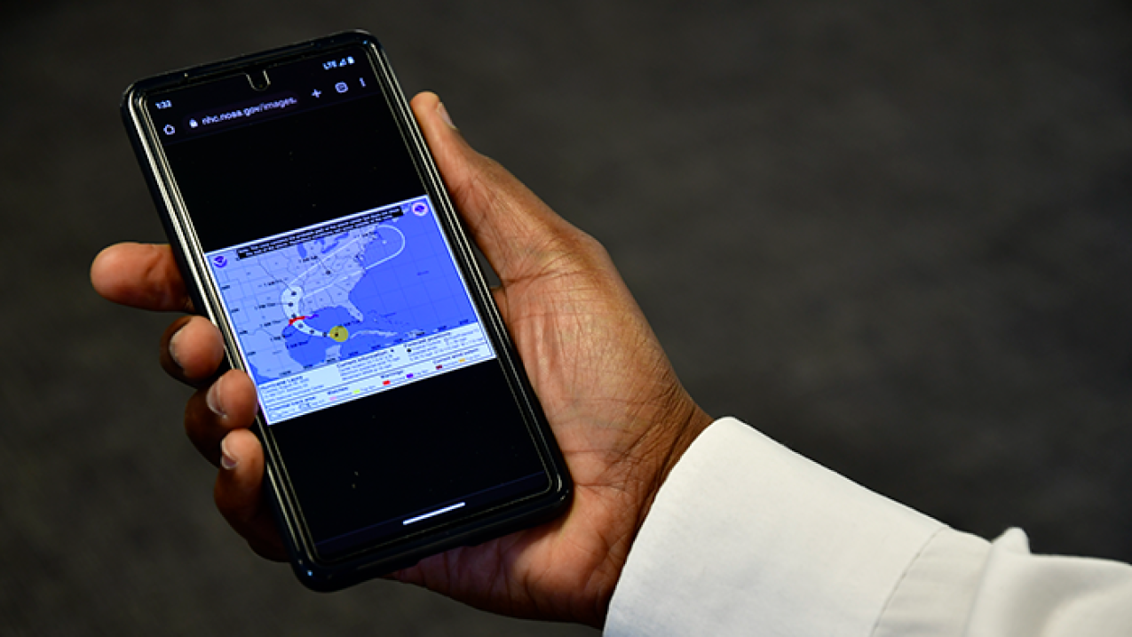 A picture of a hand holding a cell phone that is displaying a hurricane Forecast Cone graphical product.