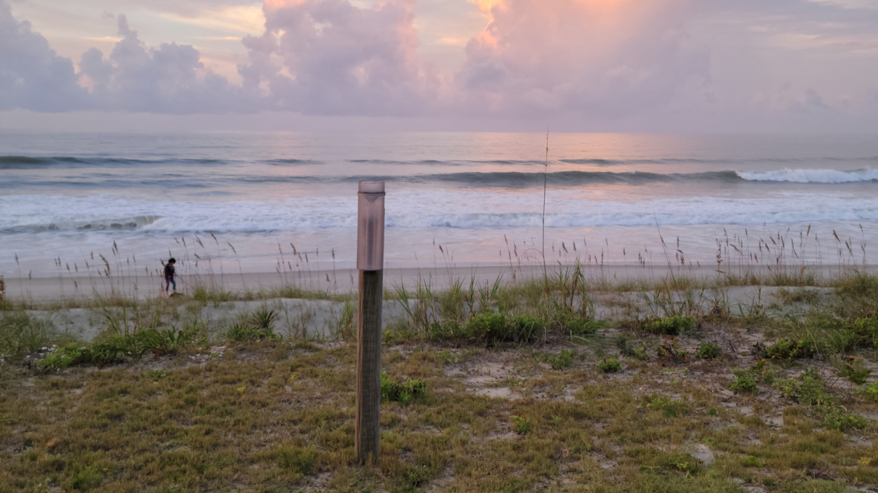 A plastic cylinder with an opening at the top to collect precipitation is mounted upright on a wooden post that stands alone in the sand dunes in front of a beach with crashing waves.