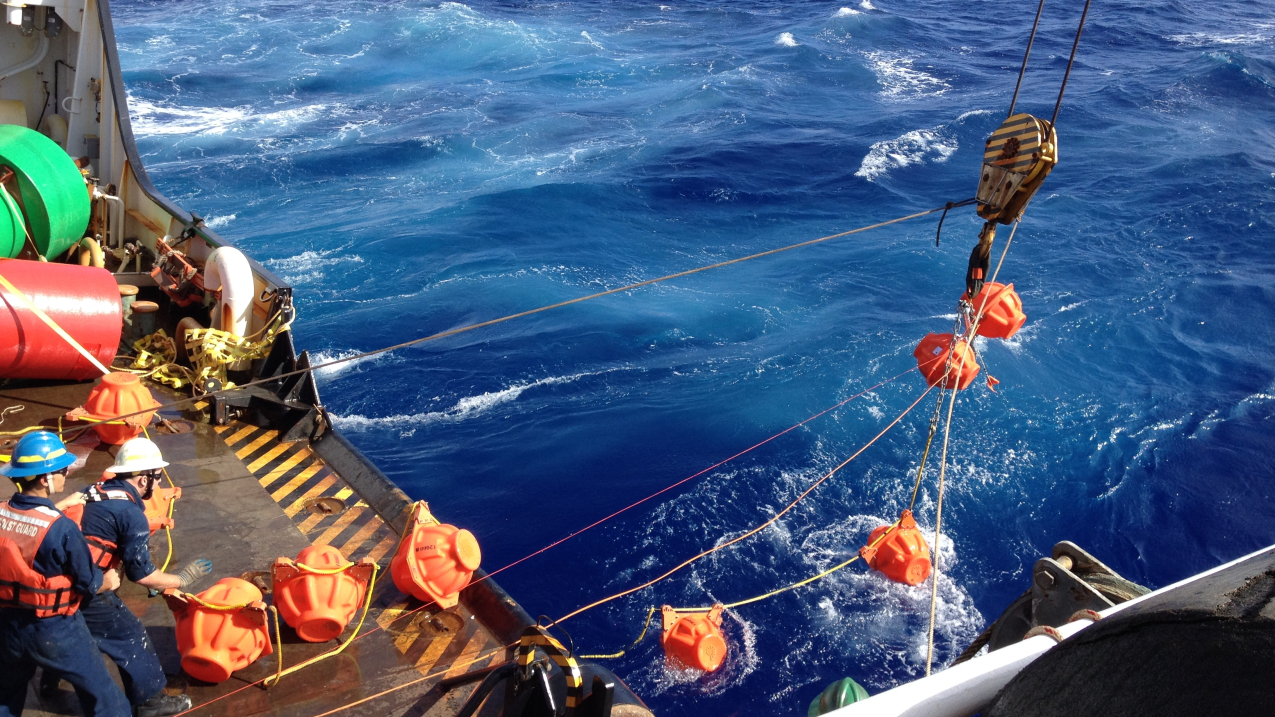 An aerial view of a ship crew recovering a hydrophone from the ocean. There are people on board the vessel with hard hats and life jackets who are using a pulley system to hoist the hydrophone out of the water.