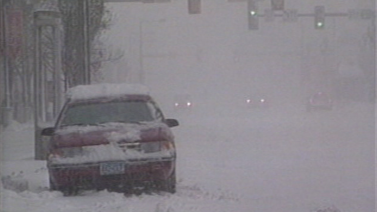 A snow-covered car sits stopped in the street during whiteout blizzard conditions. 