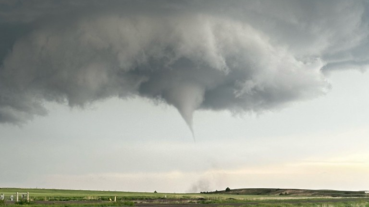 Dark clouds swirl together at the top of the frame and a tornado cone begins to form below the clouds above a cornfield. Flat farmland extends as far as the eye can see; on the horizon, the sun is showing through lighter clouds.