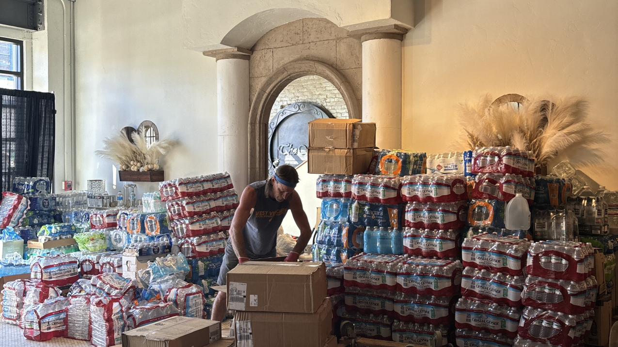 A makeshift open floor space is used as a temporary supply distribution site. In the middle of the image is a volunteer surrounded by pallets of cardboard boxes and cases of large water bottles piled 4-5 ft high. In the background is a wall and a door flanked by two pillars.