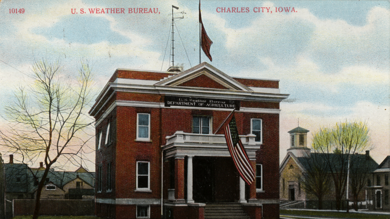 Postcard of the U.S. Weather Bureau Building at Charles City, Iowa. c. 1900. Note the Weather Forecast Flag system flags flown on the flag pole mounted on the roof. They appear to be the blue flag with a black pennant above, indicating a forecast of higher temperatures and rain.
