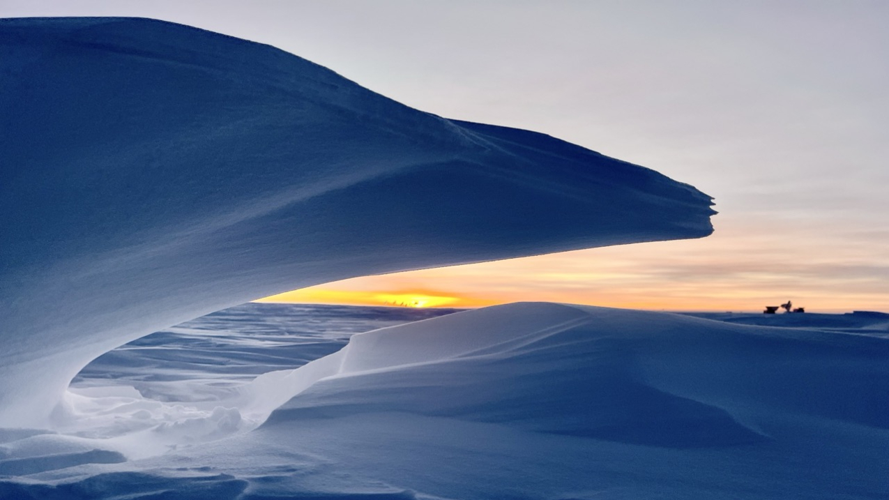 MARCH 20, 2025: The setting sun at NOAA's South Pole Atmospheric Baseline Observatory.  Dark Sector telescopes and a final glimpse of sunlight are framed by a wave of snow, setting off six months of darkness at the South Pole.