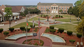 A photo of a college campus that looks down on the campus as if the photo was taken from an upper story. The campus has central lawn with decorative geometric walkways with modern fountains at the entrance. Buildings surround the central lawn.