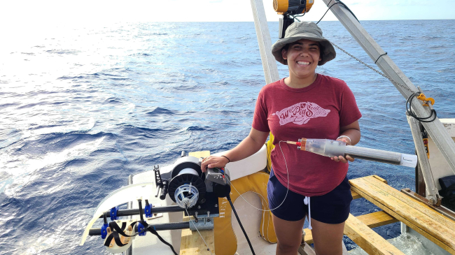 Adriana stands on a boat, holding a winch that she designed during the internship.