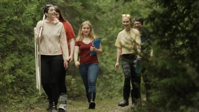 A group of students walk through a wooded area. Students carry a clipboard and other scientific data collection tools.