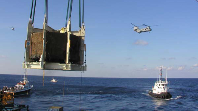 A large boom filled with a brown cargo box is hanging over the ocean while two tug boats wait in the background. A helicopter can also be seen in the sky. 
