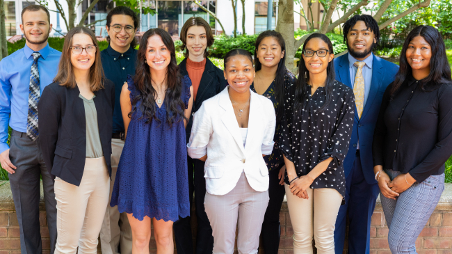 Ten young adults in business casual clothing pose outside.