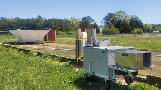 An infrared spectrometer and ceilometer sit in the foreground with a trapezoidal radar wind profiler in the background, in a grassy field in Courtland, Alabama.