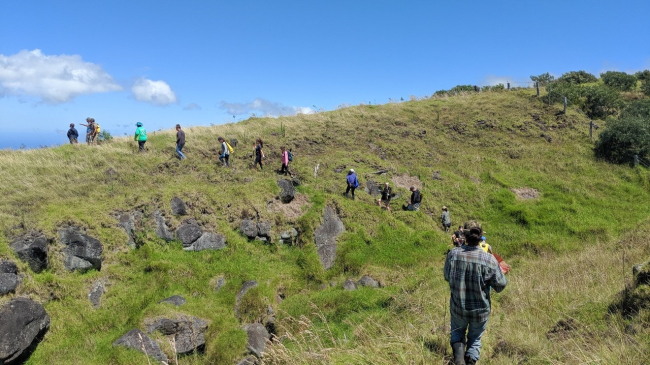 Students and educators hike along a grassy hillside in a single-file line.
