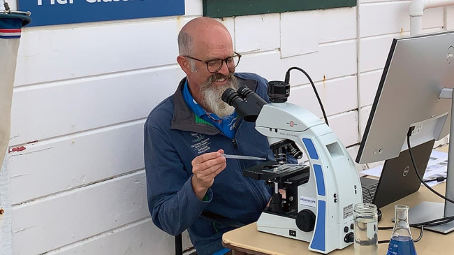 Justin Holl smiles while adding a drop of water to a slide on a microscope with a pipette. The microscope appears to be connected to a computer.