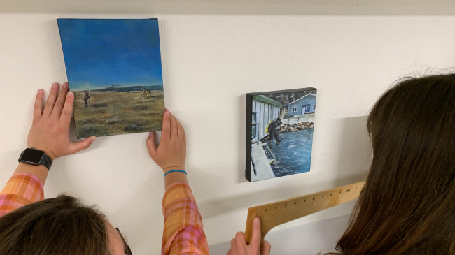 Kate holds a ruler as her sister places a painting. The left painting shows someone holding a pole that extends several feet above them while another person looks through a surveying tripod in a coastal area. The second painting shows a scuba diver mid-jump from a boat docked near the shore.