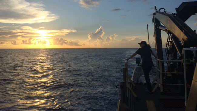 Emily Narrow, mission videographer, enjoys the sunset over the Pacific Ocean from the back deck of NOAA Ship Okeanos Explorer, during the NOAA Ocean Exploration mission: Discovering the Deep: Exploring Remote Pacific Marine Protected Areas In 2017.