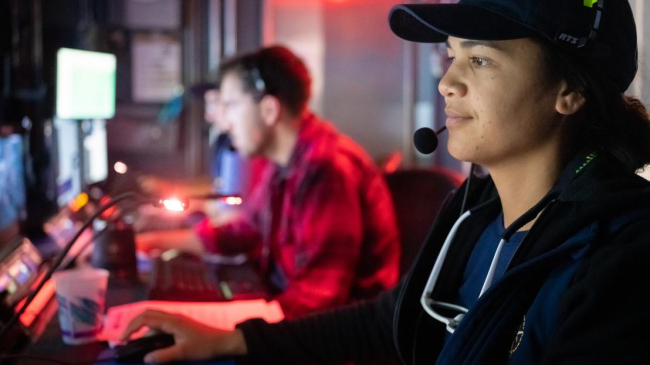 Several people sit in front of monitors wearing headsets in a room with red lights.