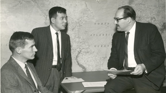 A black and white photo shows Joseph Smagorinsky, Kirk Bryan, and Suki Manabe sitting and discussing a paper in 1969.