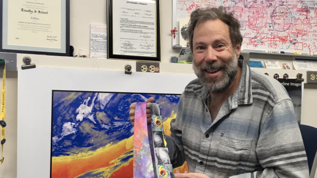 Tim Schmit shows off two brightly patterned ties in front of a photo that matches one of them.