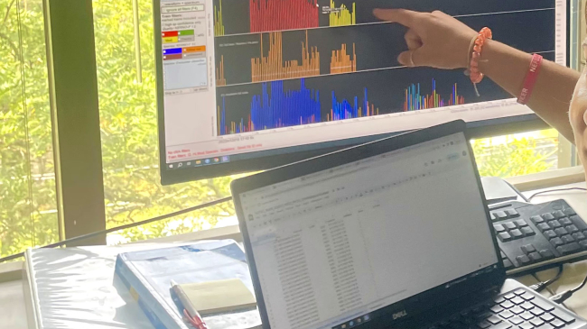 Cassidy is sitting at a work desk, looking at the camera. One of her hands rests on a laptop with data on it, while she uses her other hand to point to a computer monitor displaying multiple waveforms.