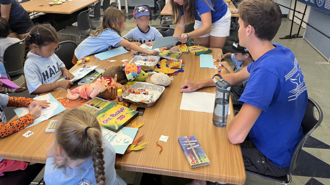 Several children are working on a craft at a table with spread out crafting supplies for coloring and pasting paper. Laura is talking to two of the students; she looks like she is explaining the craft.