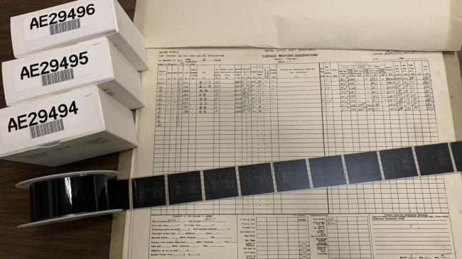 A reel of microfiche is unrolled on top of a Naval weather logbook alongside boxes of other reels.