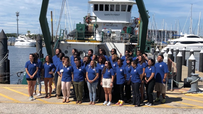 A group of people in matching blue shirts with CCME logos pose in front of a large, docked vessel.