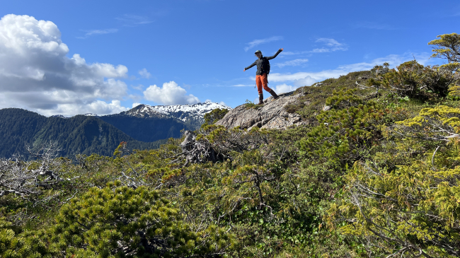 Emma Rudy posing with her arms spread on top of a mountain. The vegetation is low-lying, allowing for a clear vista of the surrounding mountains.