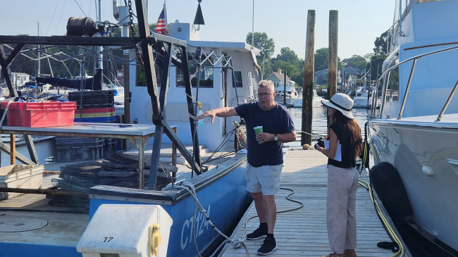 Grace and the oyster farmer stand on a dock with boats docked on either side. Grace looks at the oyster grower, who is speaking and gesturing towards his boat.