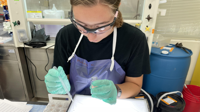 Makayla stands at a lab bench, looking down at a lab tray that looks to contain shallow water. She holds a transfer pipette in one gloved hand and a sampling vial in the other. She wears protective goggles and an apron.